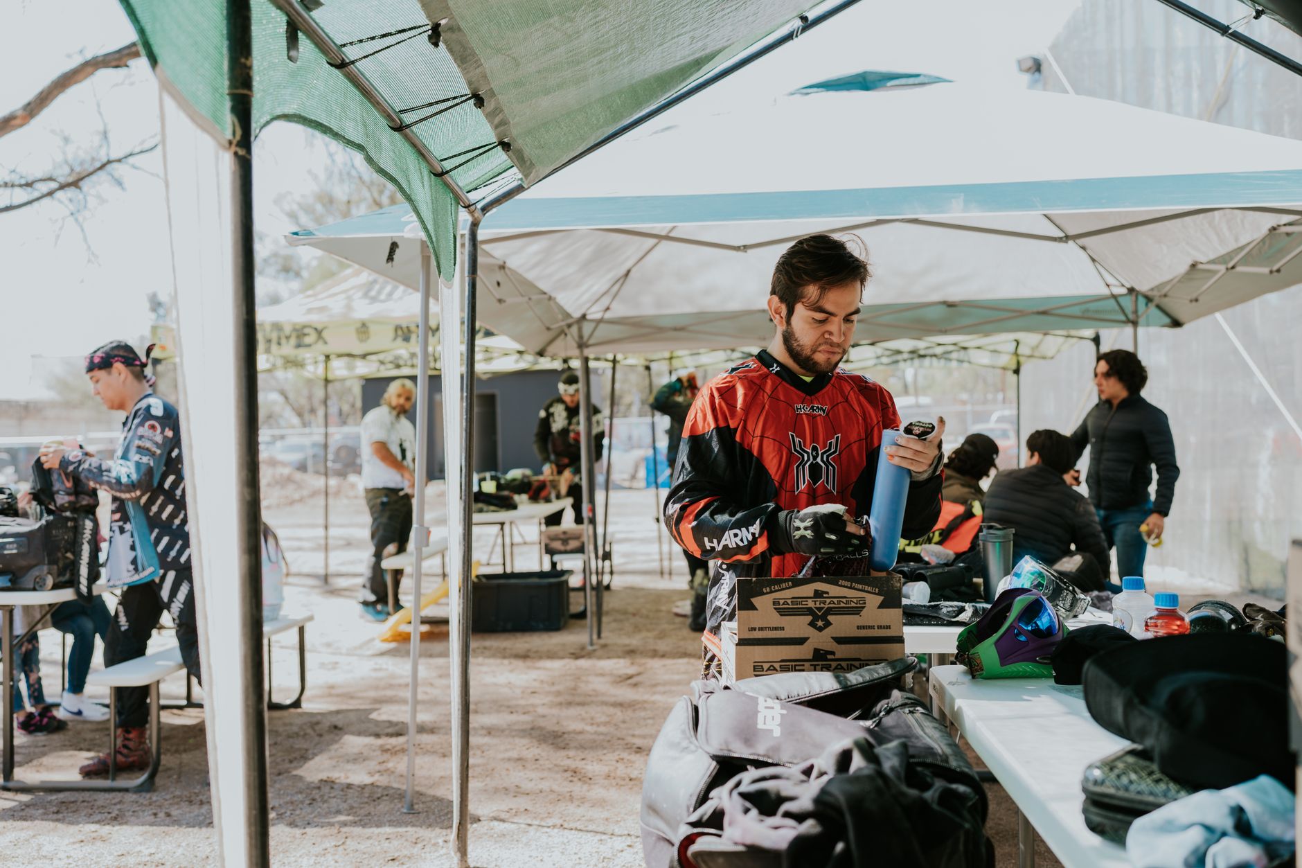 Group of adult men preparing gear for paintball under outdoor tents in daytime.