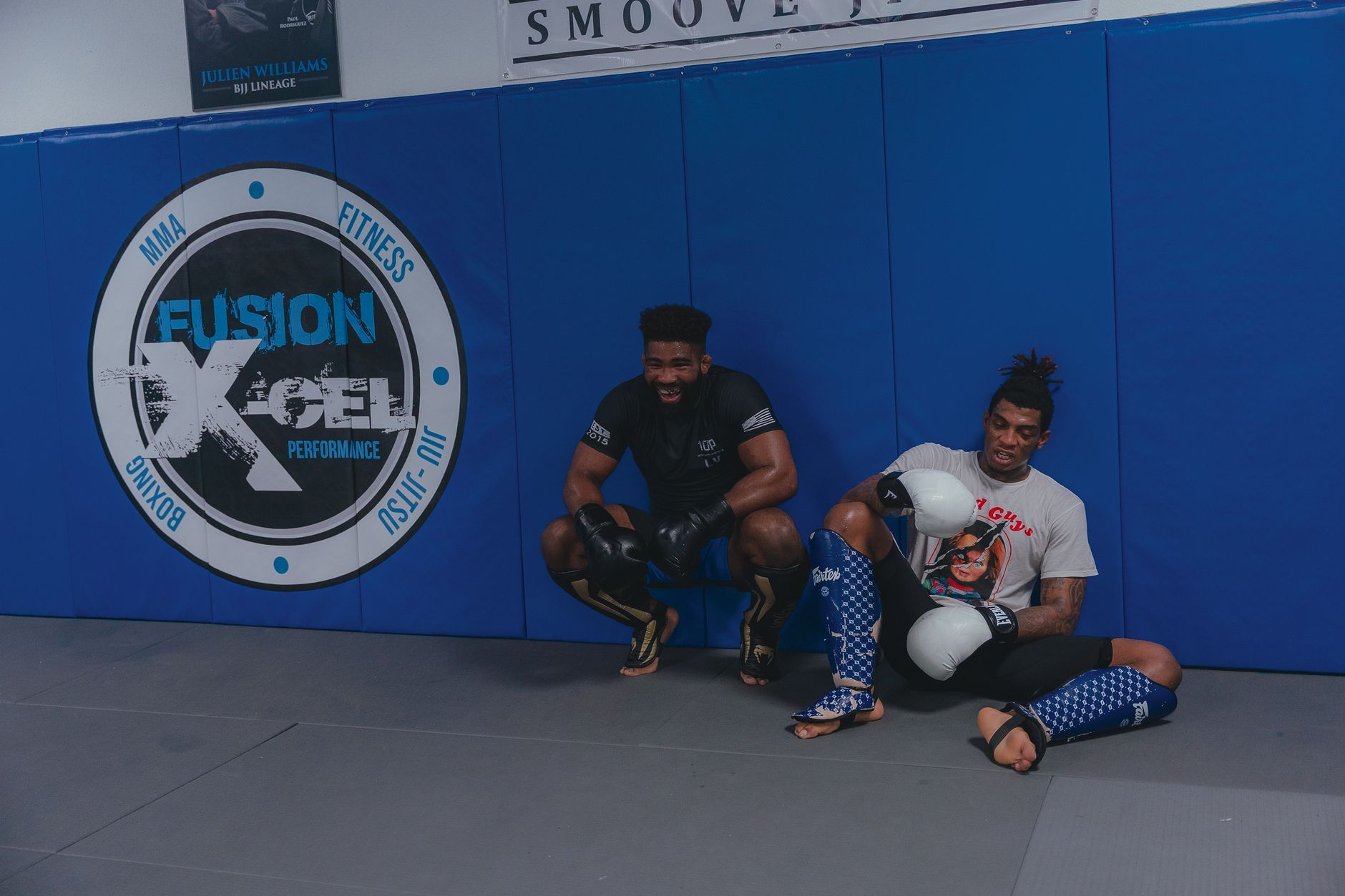 Two young men sitting and resting in an MMA gym, wearing boxing gloves and gear, smiling.