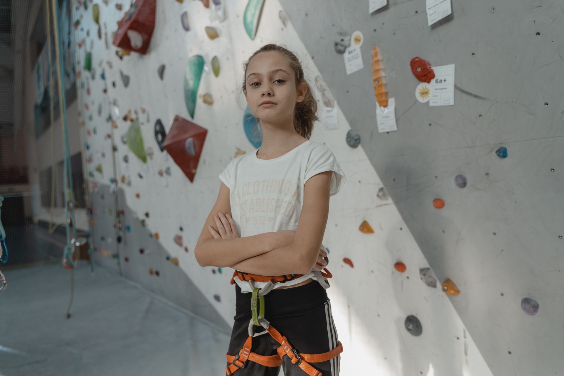 Young girl with crossed arms poses confidently in an indoor climbing gym wearing a harness.