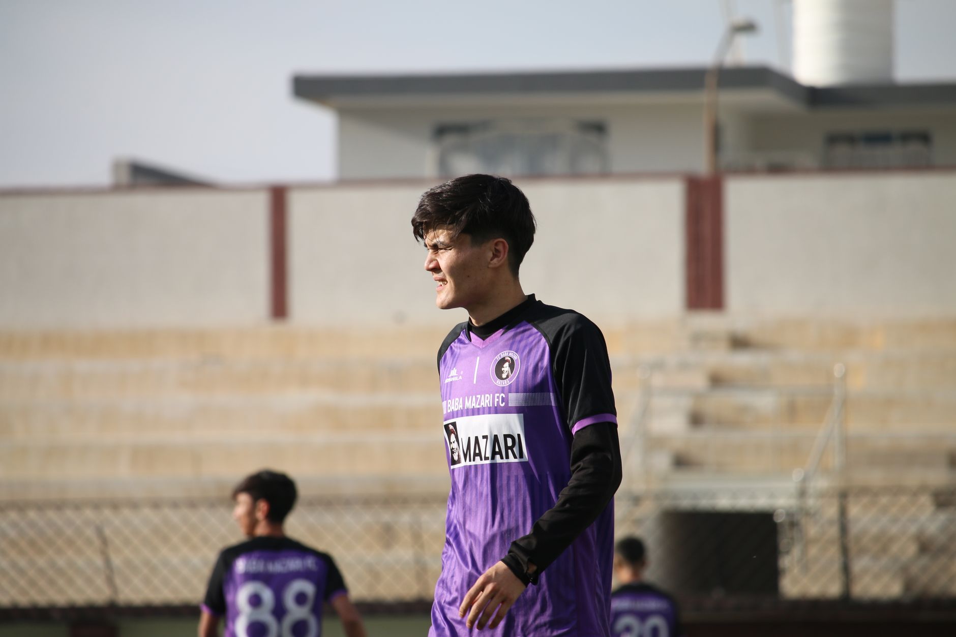 A young male soccer player wearing a purple jersey stands on an outdoor field.