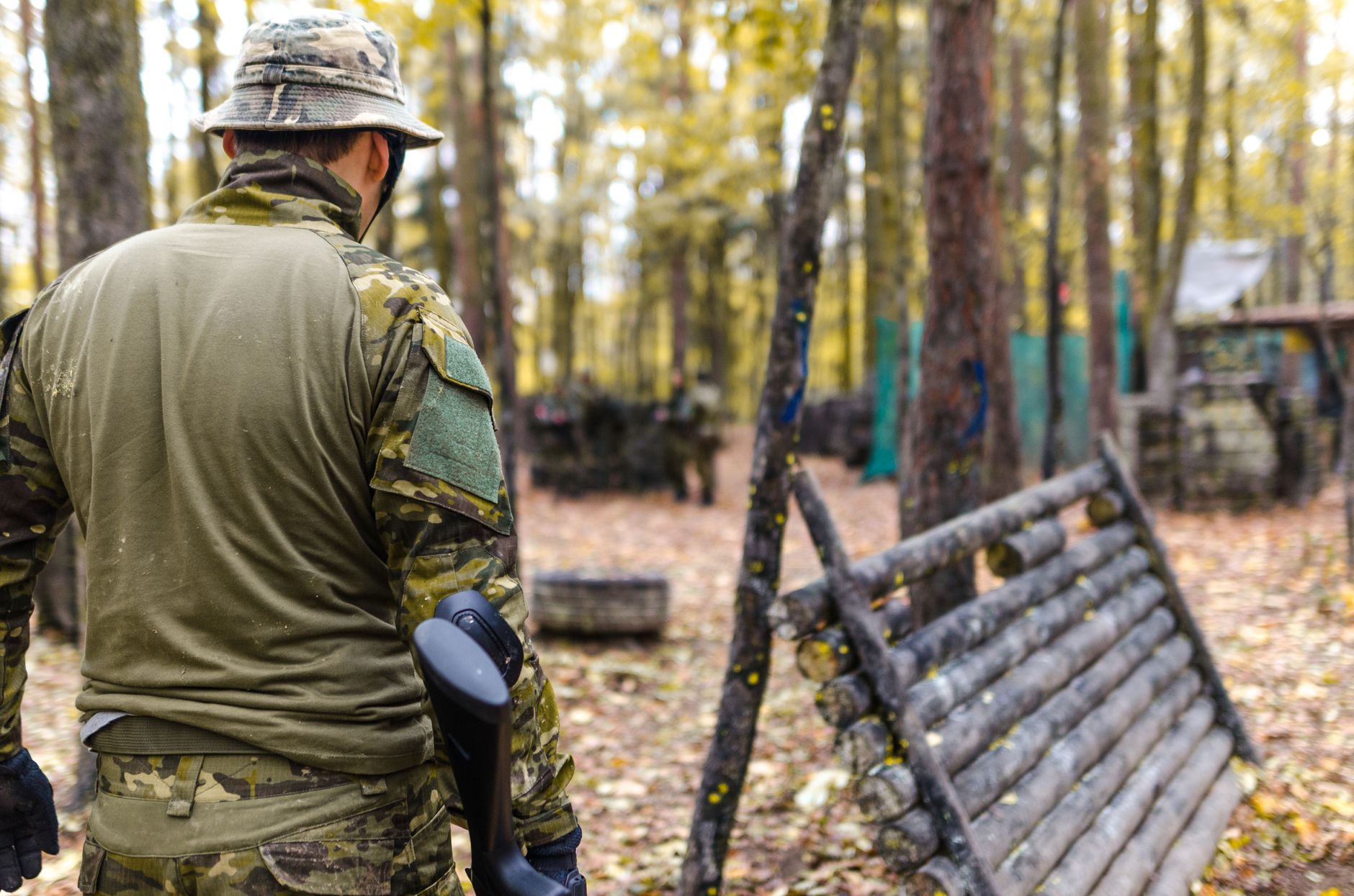 A man wearing camouflage gear participates in an outdoor paintball game in a forested area.