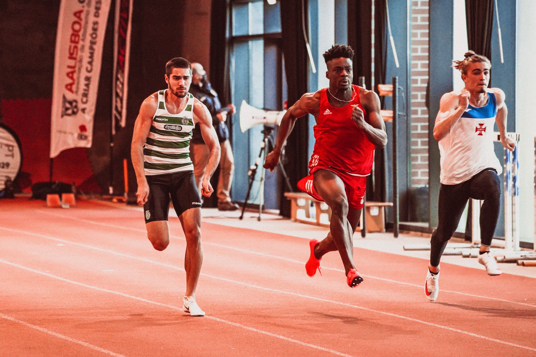 Three athletes sprinting on an indoor track during a sports competition.