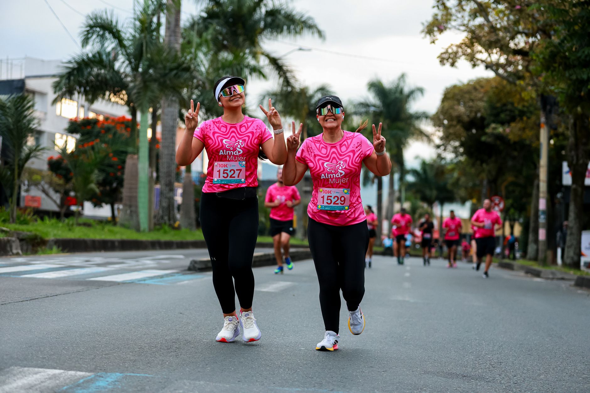 Two women in pink shirts running a charity race outdoors, showing peace signs.