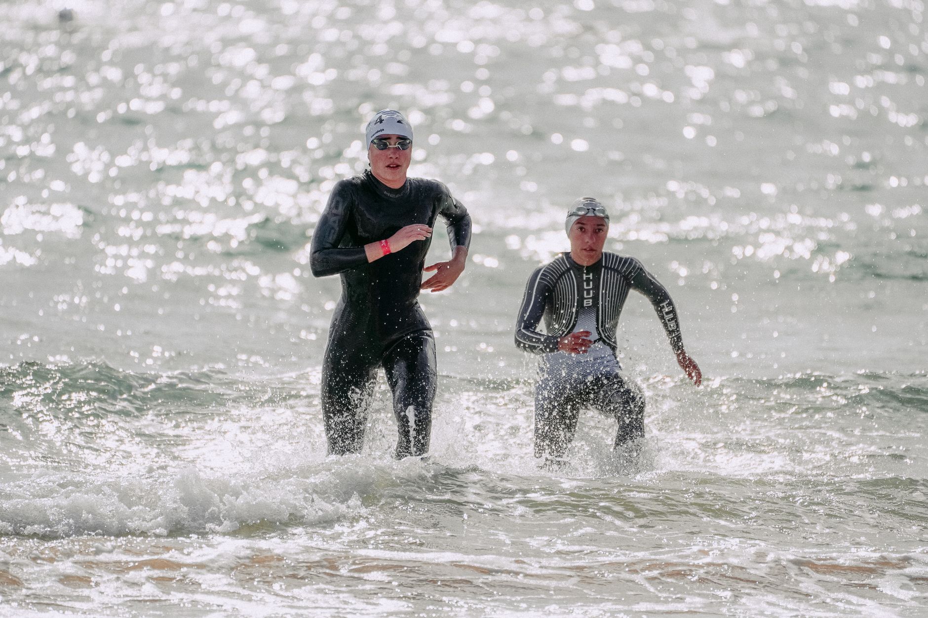 Focused young people in wet suits and swim caps running out of wavy glistening sea in sunlight