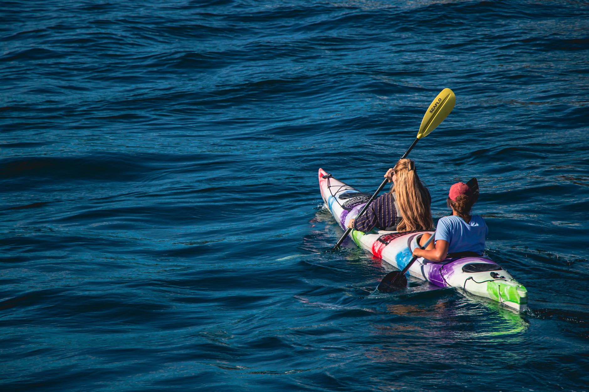 Two women paddle a colorful kayak on a calm lake during a sunny summer day, enjoying outdoor recreation.