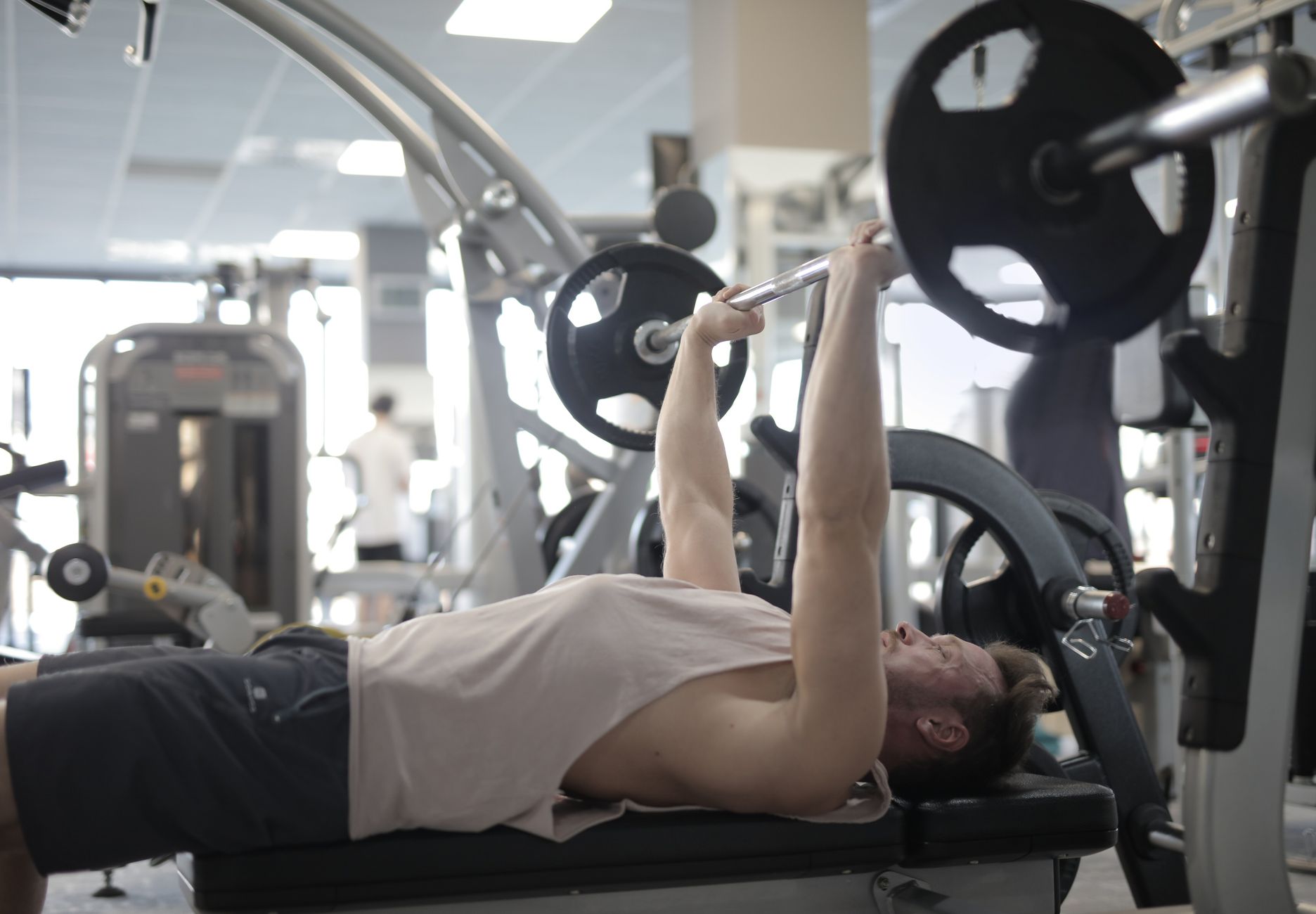 Side view of strong young sportsman in casual sportswear pressing heavy barbell on bench while intense training in modern fitness center