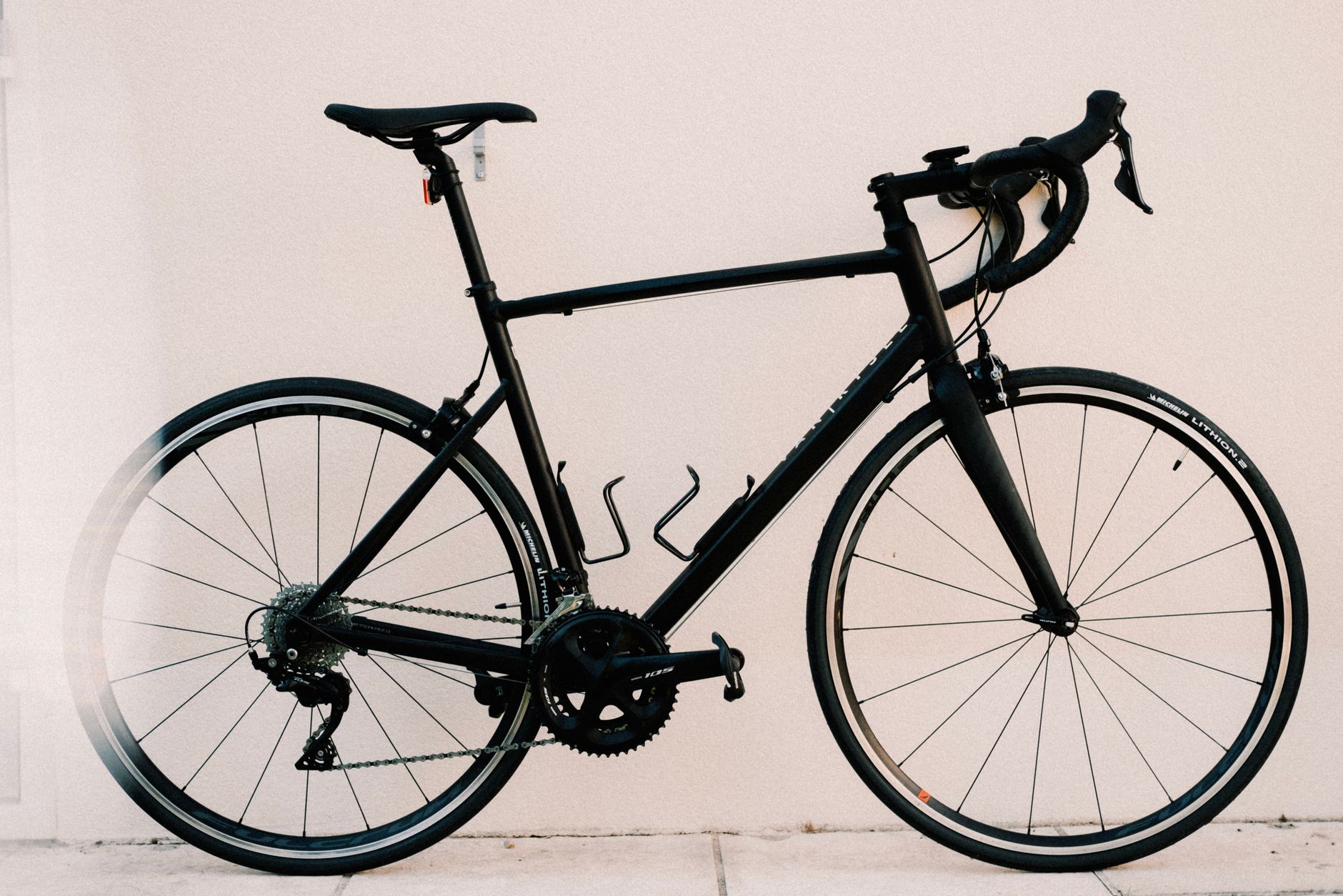 Black road bike with drop handlebars leaning against a beige wall.