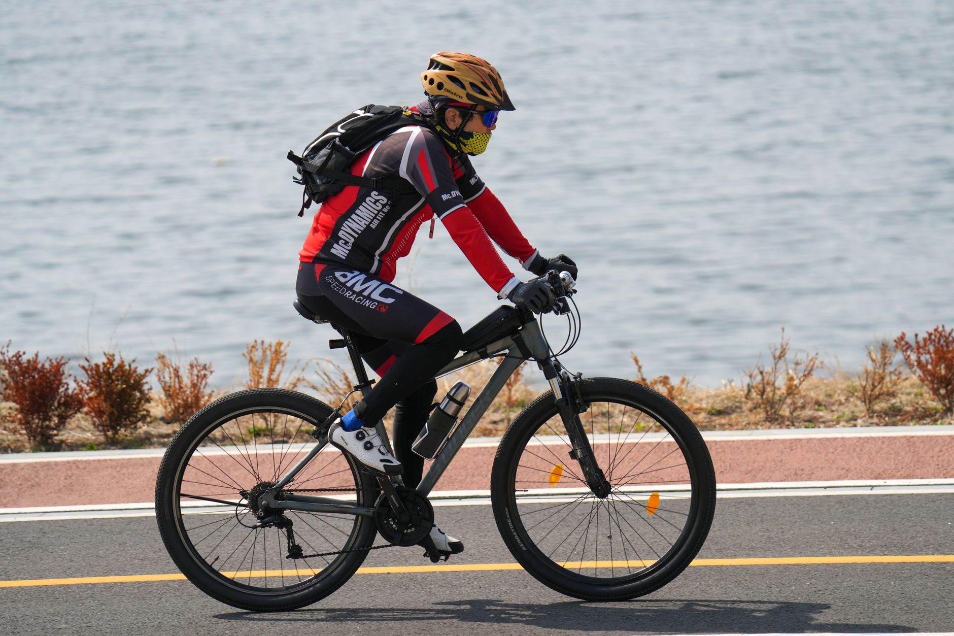 Cyclist wearing safety gear rides along a waterfront on a sunny day, showcasing healthy outdoor activity.