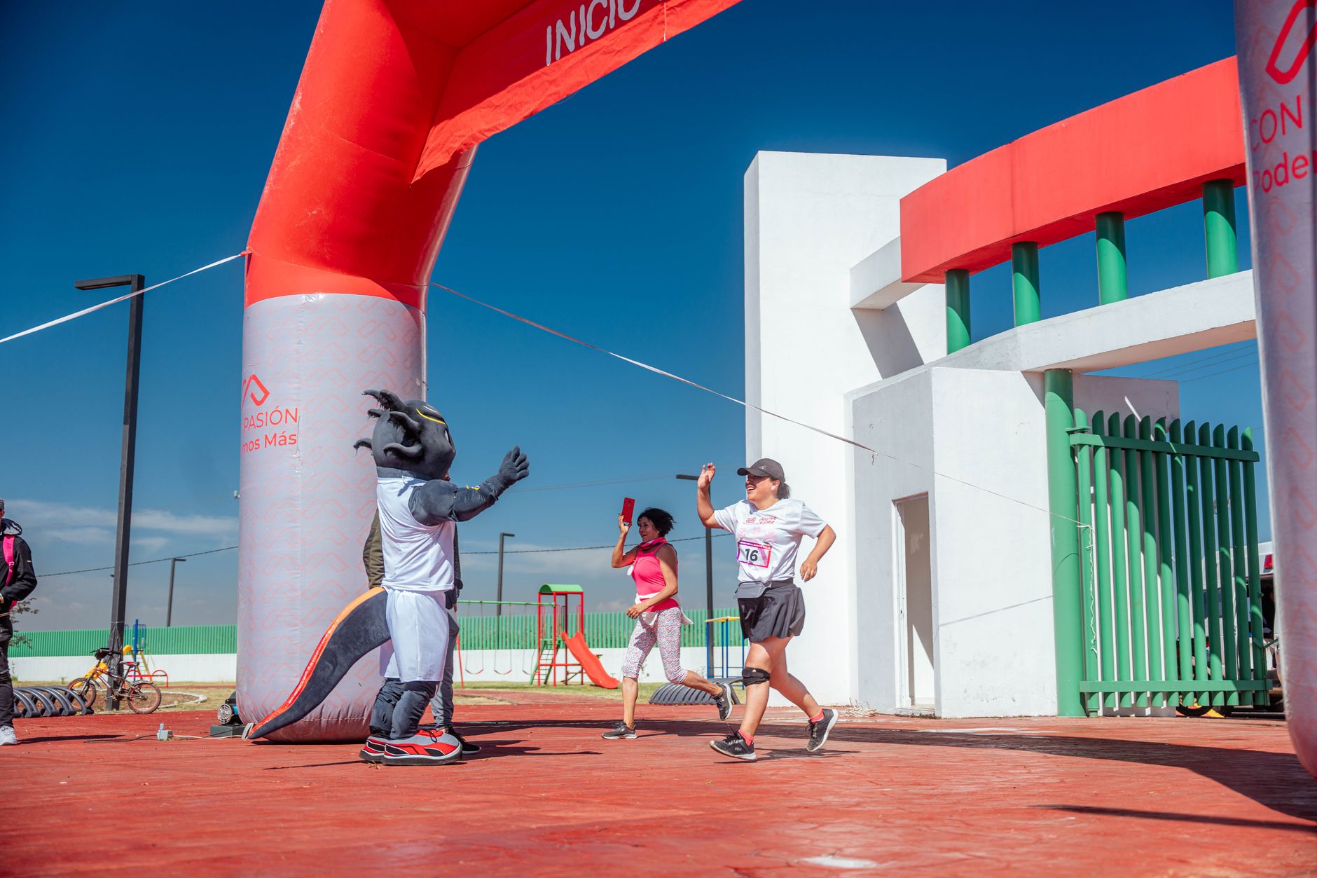 Runners cross the finish line under a colorful arch with a cheering mascot during a sunny outdoor event.