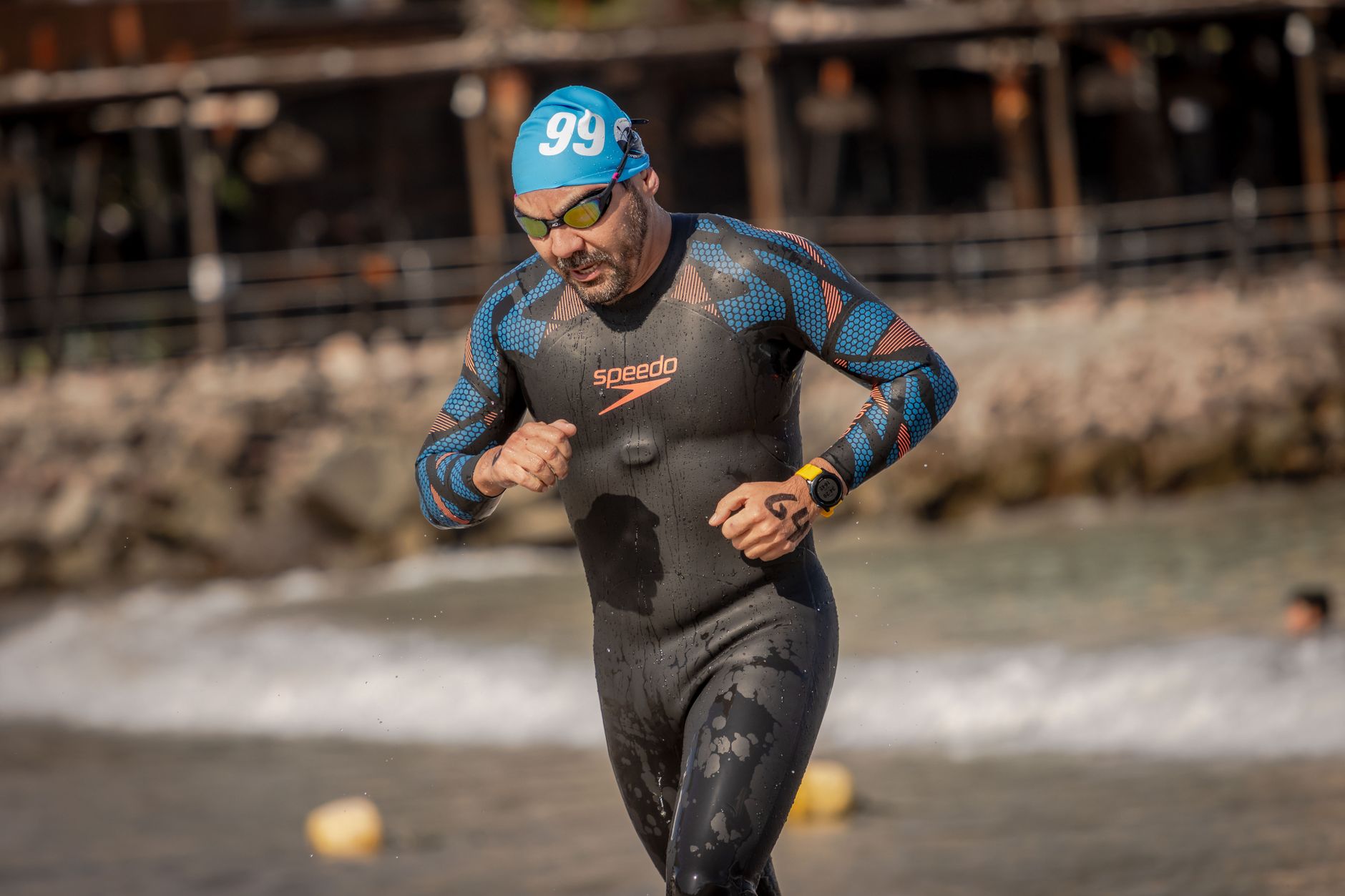 Male athlete runs on the beach after a swim, wearing a wetsuit and swim cap.