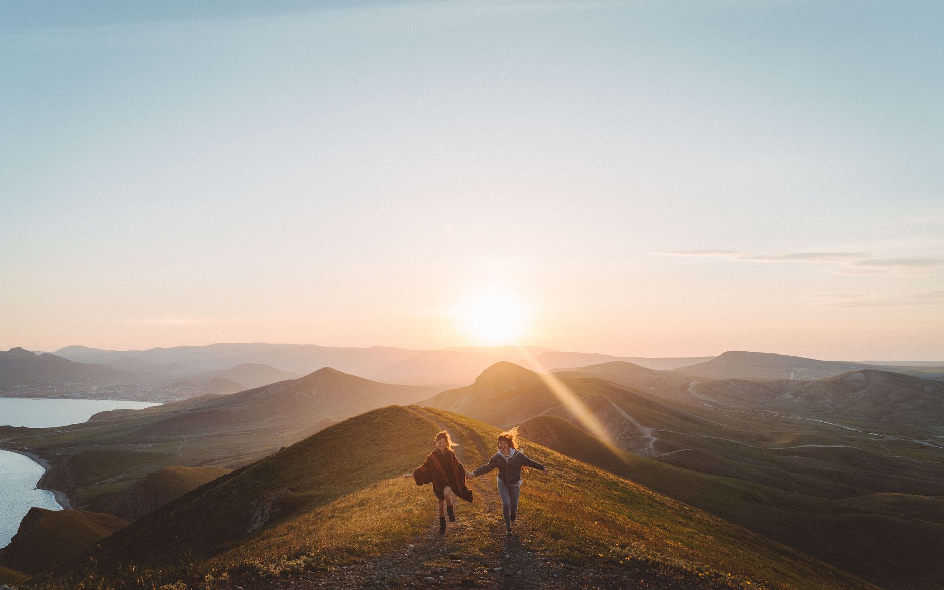 Two women running hand-in-hand down a hilltop trail at sunset with a stunning mountainous landscape backdrop.