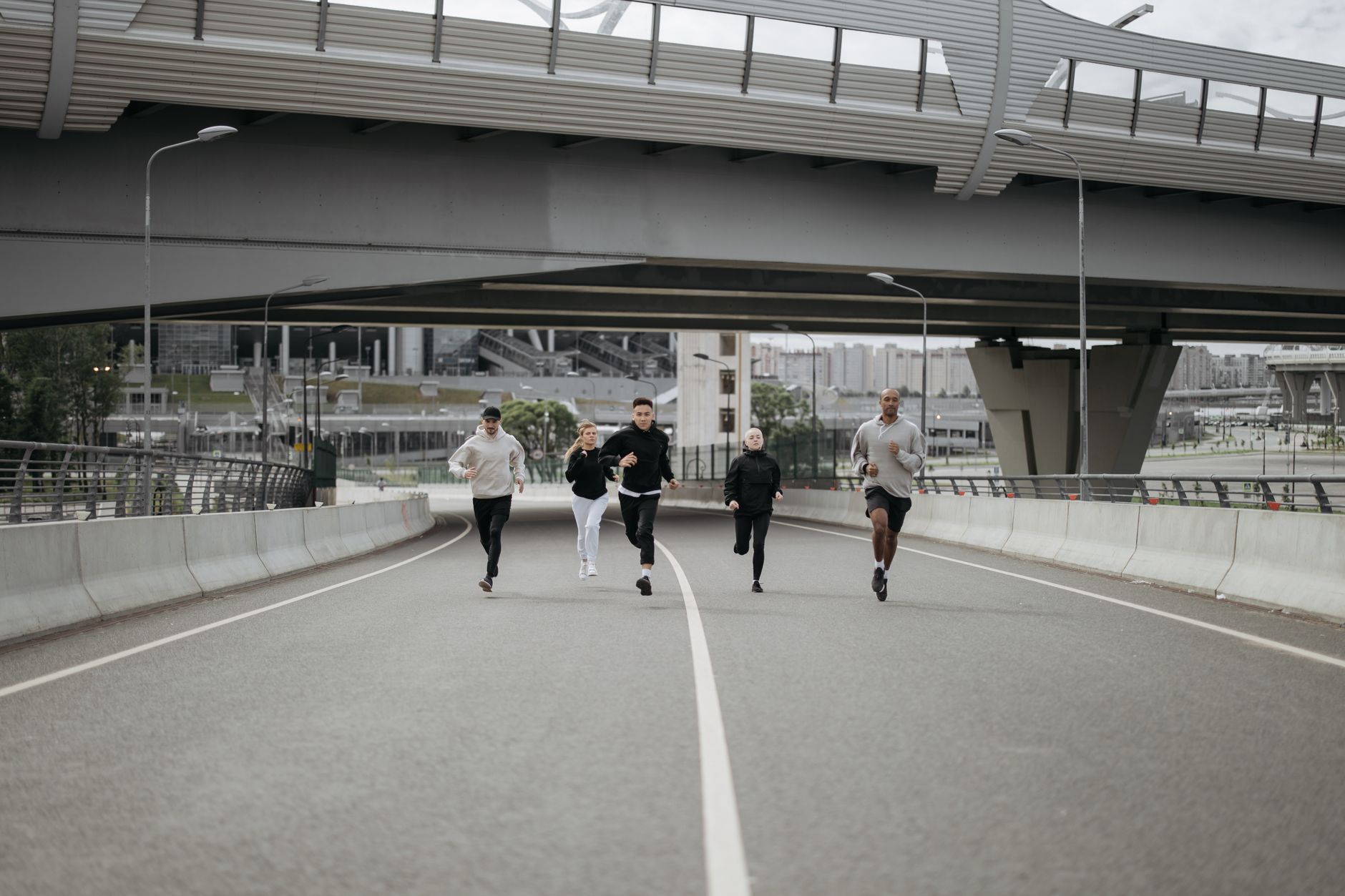 A diverse group of people running on a city road, showcasing teamwork and fitness under an overpass.