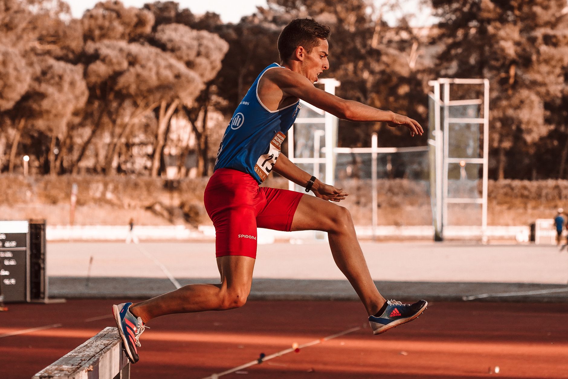 Focused athlete in blue tank top and red shorts leaping hurdle on outdoor track.