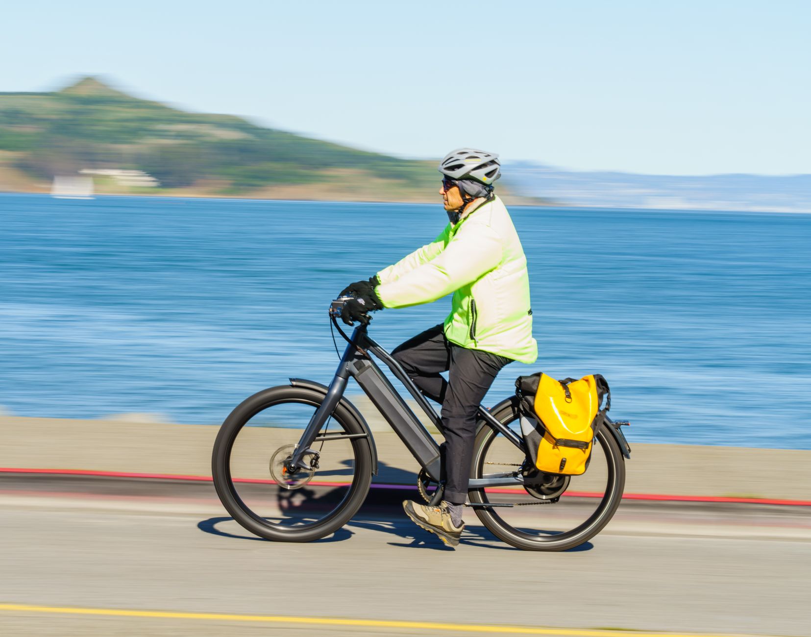 A person in neon gear cycles an e-bike with a yellow pannier on a seaside road.